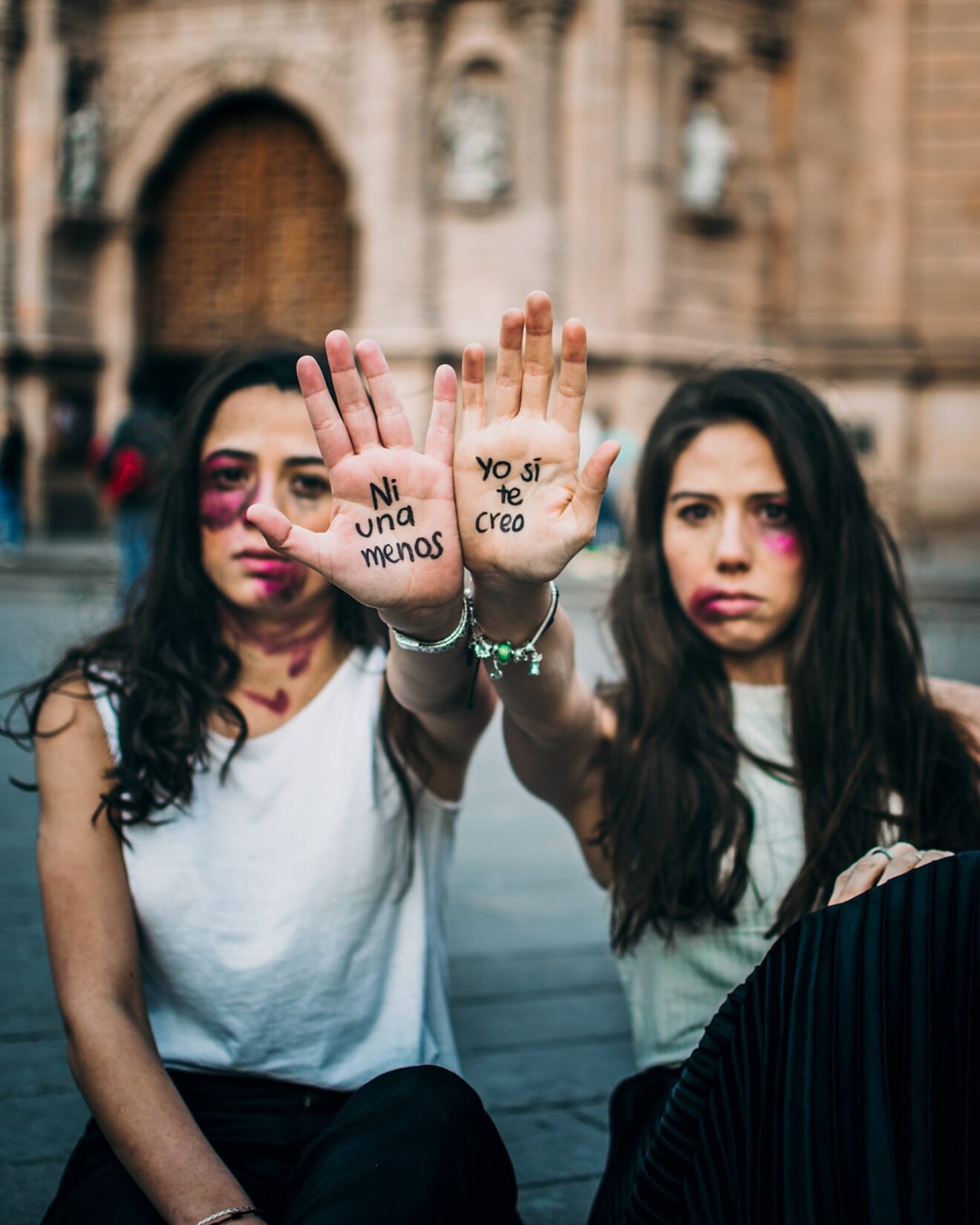 Two women sit on the ground, each with one hand up, palm facing the camera.  On their palms there is writing that says "Ni una menos" and "Yo si te creo".  They both also have makeup on their face made to look like bruises on their mouths and eyes.